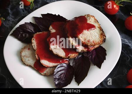 Steak juteux rôti de porc bien cuit avec sauce rouge et feuilles de basilic sur une assiette blanche sur une table en marbre noir. Tomates cerises et brins de basilic sur un mar Banque D'Images