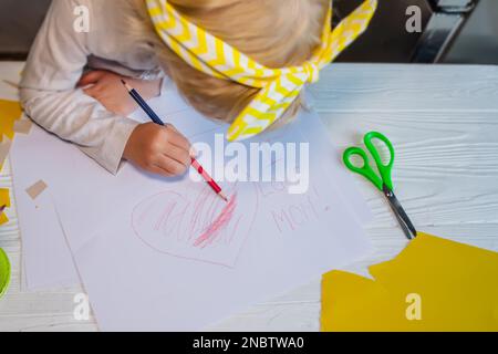 Une petite jeune fille peint un coeur peint sur une carte postale pour la fête des mères. Petite fille écrit félicitations pour maman Banque D'Images