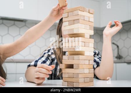 Les écoliers jouent à jenga assis près de la table de cuisine. Main de fille a mis un petit bloc de bois sur le dessus de la tour en gardant l'équilibre. Les amis se détendent à la maison Banque D'Images