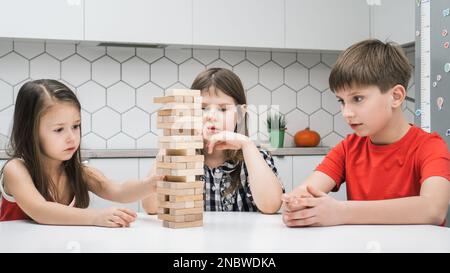 Les enfants de l'école concentrée jouent tour assis à table de cuisine. Garçons et filles construisent tour à partir de petits blocs de bois. Les amis se détendent à la maison, en période de jeu Banque D'Images