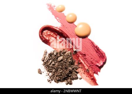 Nuances de maquillage : brun foncé, rouge à lèvres rouge et rose et gouttes de crème de couleur peau sur fond blanc. Banque D'Images