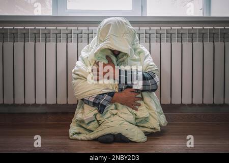 Il fait froid à la maison en hiver. L'homme gèle dans sa maison en hiver à cause d'un thermostat cassé. Triste jeune gars enveloppé dans des frissons de plaid pendant que stit Banque D'Images