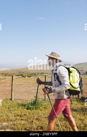 Vue latérale de l'homme senior afro-américain portant un chapeau avec sac à dos et des bâtons de randonnée sur terre. Espace de copie, inchangé, ciel clair, retraite, vacances, s Banque D'Images