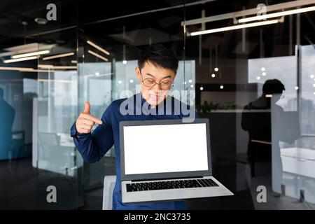 Inquiet et contrarié jeune asiatique homme, programmeur, homme d'affaires, indépendant debout dans le bureau et tenant un ordinateur portable cassé. Pointer un doigt en colère sur un écran vierge éclairé. Banque D'Images