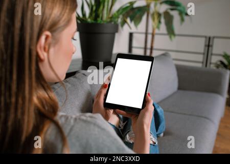 Une jeune femme blonde décontractée et méconnue dans un salon moderne, posée sur un canapé confortable à la maison, travaillant avec une maquette de tablette à écran vierge Banque D'Images