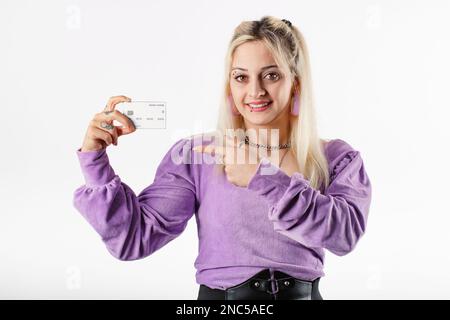 Blonde gaie femme portant un chemisier à côtes lilas isolé sur fond blanc tient une carte de crédit dans la main et des points à la carte de débit avec l'index de la finge Banque D'Images