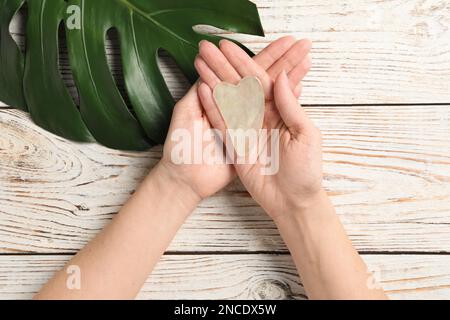 Femme tenant l'outil gua sha sur une table en bois blanc, vue de dessus Banque D'Images