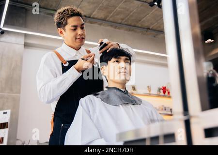 Joyeux coiffeur de jeune asiatique souriant portant un tablier dans moderne salon tenant la brosse et les ciseaux de couper les cheveux de client Banque D'Images