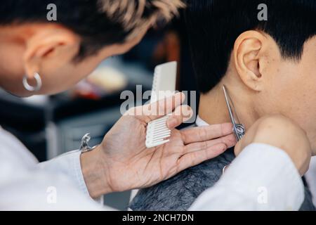 Joyeux coiffeur de jeune asiatique souriant portant un tablier dans moderne salon tenant la brosse et les ciseaux de couper les cheveux de client Banque D'Images