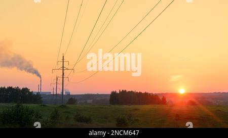 Piliers de lignes électriques avec des fils se tenant dans une rangée à l'extérieur de la centrale thermique de la ville avec des cheminées fumeurs au coucher du soleil dans un champ sur l'herbe verte Banque D'Images