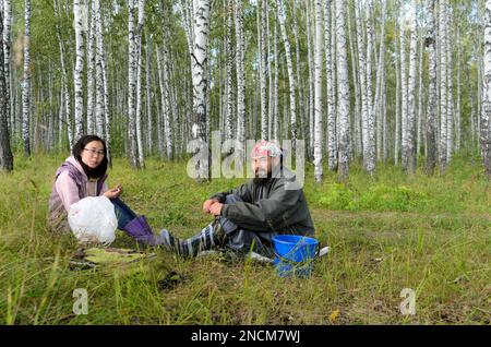 Homme et fille souriant assis avec soin dans l'herbe dans un champ près d'une forêt de bouleaux reposant avec un seau et un paquet à Sibir . Banque D'Images