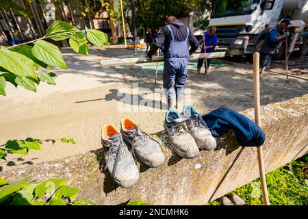 Vieilles chaussures de sécurité usées doublées, placées sur un petit mur près du chantier de construction, car les ouvriers des bottes utilisent un râteau pour épandre du béton frais Banque D'Images