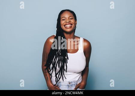 Une jeune femme heureuse souriant à l'appareil photo tout en se tenant sur un fond bleu dans des vêtements décontractés. Jeune femme noire élégante portant des tatouages de bras et Banque D'Images