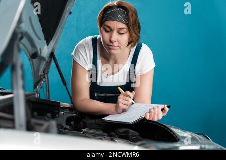 Le portrait d'une jeune femme de mécanicien caucasien en combinaison signe des documents au service de voiture. Le concept de l'égalité de la femme et travaille à la réparation automatique s. Banque D'Images