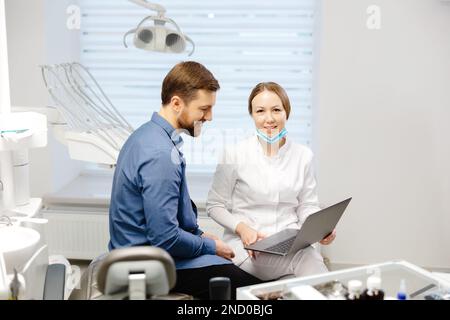 Un beau jeune homme parle à une femme médecin à un rendez-vous dentaire dans un beau bureau lumineux. Le dentiste explique au patient et montre Banque D'Images