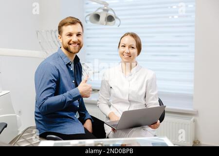 Un beau jeune homme parle à une femme médecin à un rendez-vous dentaire dans un beau bureau lumineux. Le dentiste explique au patient et montre Banque D'Images