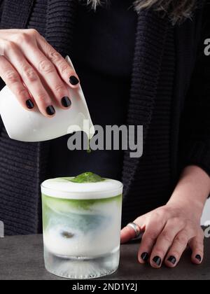 Une femme anonyme verse une boisson aromatique au matcha dans une tasse de verre avec du lait et du cube de glace tout en préparant une boisson saine Banque D'Images