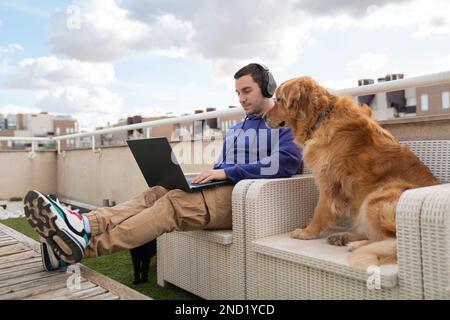 Jeune homme positif indépendant dans des vêtements décontractés et des écouteurs assis sur un banc tout en travaillant à distance via un ordinateur portable près de mignon Golden Retriever chien duri Banque D'Images