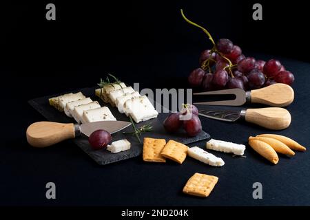 Vue de dessus d'un assortiment de délicieux fromages disposés sur un tableau noir avec des raisins mûrs et des craquelins placés près des couteaux sur une surface sombre Banque D'Images