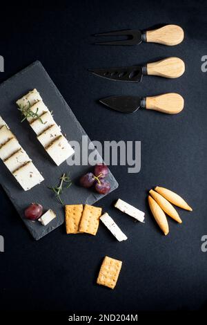 Vue de dessus d'un assortiment de délicieux fromages disposés sur un tableau noir avec des raisins mûrs et des craquelins placés près des couteaux sur une surface sombre Banque D'Images