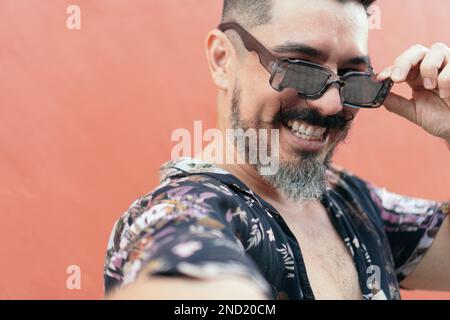 Positif barbu mâle hipster dans des lunettes de soleil et chemise ornementale souriant largement tout en regardant l'appareil photo contre le mur orange Banque D'Images