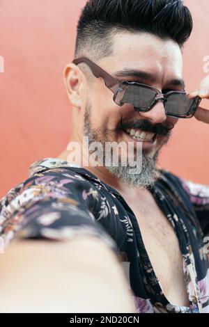 Positif barbu mâle hipster dans des lunettes de soleil et chemise ornementale souriant largement tout en regardant l'appareil photo contre le mur orange Banque D'Images