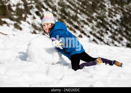 Vue latérale complète de la fille positive en vêtements d'extérieur et chapeau faisant boule de neige tout en faisant bonhomme de neige sur le sol enneigé en forêt d'hiver Banque D'Images