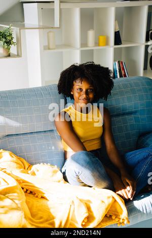 Contenu afro-américaine jeune femme avec une coiffure afro en tenue décontractée reposant sur un canapé confortable regardant l'appareil photo à la maison Banque D'Images