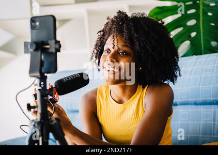Jeune Afro-américaine positive dans des vêtements décontractés avec une coiffure Afro souriant et regardant un smartphone tout en ayant la vidéoconférence sur le canapé W. Banque D'Images