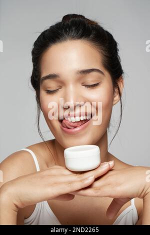 Une jeune femme ravie en haut blanc souriant tout en tenant une bouteille de produit cosmétique Banque D'Images