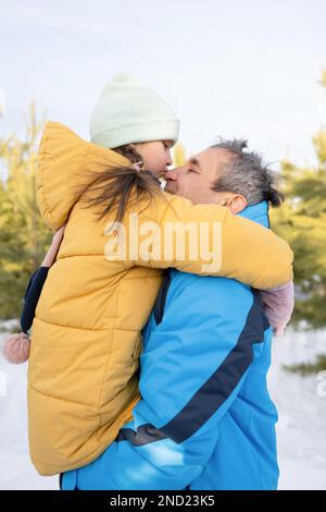 Vue latérale d'une adorable préadolescente qui embrasse son père tout en se tenant ensemble sur une pelouse enneigée dans des vêtements chauds pendant le week-end à la campagne Banque D'Images