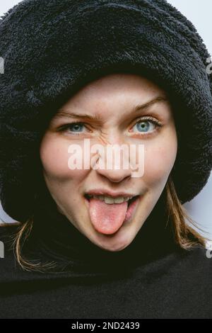 Portrait de jeune femme drôle en col de cygne noir et chapeau regardant la caméra tout en montrant la langue et exprimant des émotions sur fond blanc Banque D'Images
