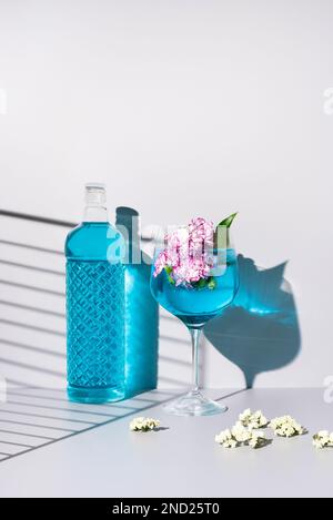 Bouteille en verre de liquide bleu placée près du verre avec cocktail rafraîchissant avec glace et fleurs sur table sur fond blanc Banque D'Images