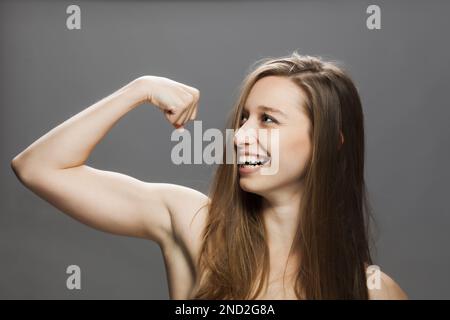 Petite fille de brunette moulante montrant des muscles et souriant. Portrait de studio sur fond gris. Banque D'Images