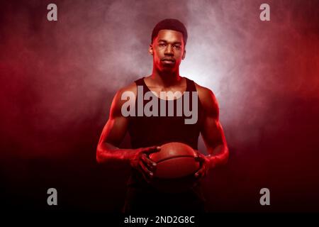 Côté joueur de basket-ball éclairé de couleur rouge tenant une balle sur fond de fumée. Sérieux homme afro-américain concentré. Banque D'Images