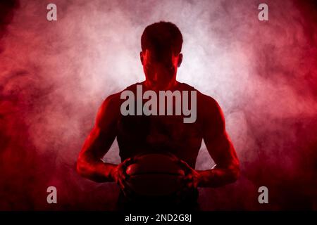 Côté joueur de basket-ball éclairé de couleur rouge tenant une balle sur fond de fumée. Sérieux homme afro-américain concentré. Banque D'Images