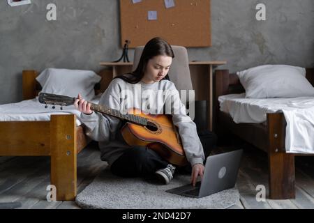 Une adolescente asiatique sérieuse apprend à jouer de la guitare à l'aide d'un ordinateur portable assis près du lit à la maison. Banque D'Images