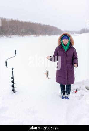 Une fille asiatique sourit et se réjouit sur la glace de la rivière en hiver en tenant un poisson pêché perchoir sur une ligne de pêche à côté de la foreuse de glace et de poisson Banque D'Images