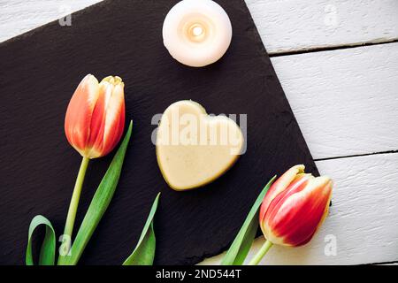 Crème pour les mains hydratante solide maison. Bar à crème en forme de coeur fait de tous les ingrédients naturels. Fond de plateau en pierre noire avec tulipes fraîches. Banque D'Images