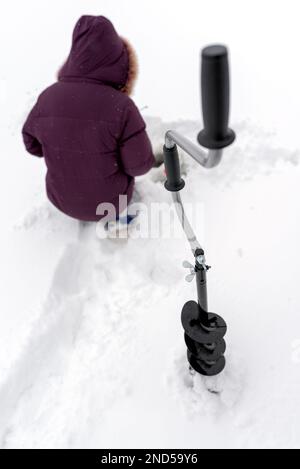 Exercice de glace pour la pêche d'hiver avec des vis se tient devant la fille assise et des trous de pêche sur la glace de la rivière dans la neige blanche. Banque D'Images