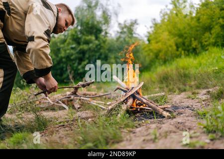 Vue à angle bas d'un pêcheur qualifié mettant du bois de chauffage sur feu de camp à faire feu sur la rive de la rivière avant de cuisiner le soir avant le coucher du soleil. Banque D'Images