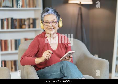 La femme âgée est heureuse de lui envoyer des SMS tout en portant des lunettes roses et des écouteurs jaunes à la maison, reflétant un style de vie détendu et moderne avec un accent sur la technologie et la communication. Photo de haute qualité Banque D'Images