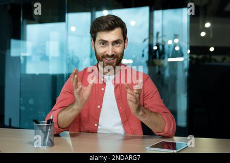 Un jeune enseignant de sexe masculin enseigne en ligne, donne des conférences, donne des leçons. Assis dans le bureau à la table devant la caméra. Parle lors d'un appel vidéo, explique, sourit. Banque D'Images