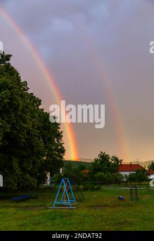 Un cliché vertical du bel arc-en-ciel brillant dans le ciel sombre au-dessus de l'arrière-cour avec une balançoire Banque D'Images