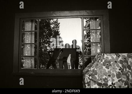 Selfie monochrome de trois personnes réfléchi dans une fenêtre dans un chalet d'été. Avec un barbecue couvert dans le coin. Juillet 1977 Banque D'Images