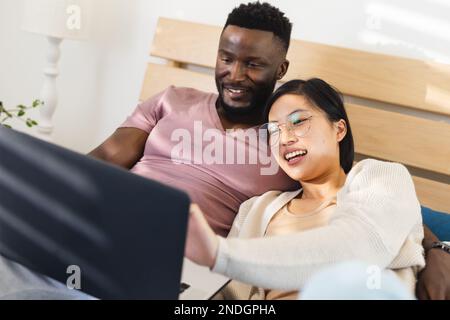 Joyeux couple varié allongé dans le lit et utilisant un ordinateur portable. Passer du temps de qualité à la maison, la vie domestique et le concept de style de vie. Banque D'Images
