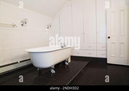 Baignoire à griffes en fonte antique blanche sur plate-forme surélevée en granit noir dans la salle de bains principale avec armoires et tiroirs blancs dans la vieille maison de 1900. Banque D'Images