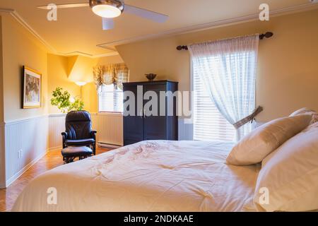 Grand lit, armoire en bois et fauteuil inclinable en cuir dans la chambre principale à l'étage à l'intérieur d'une maison de style chalet. Banque D'Images