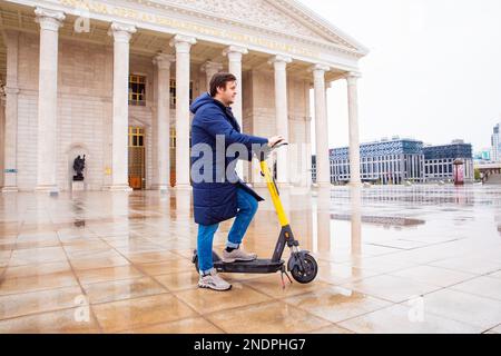 Homme et scooter électrique avec le panneau jaune Whoosh garés sur la place humide de l'opéra. Opéra et théâtre de ballet d'Astana. Partage de scooter électrique. Banque D'Images