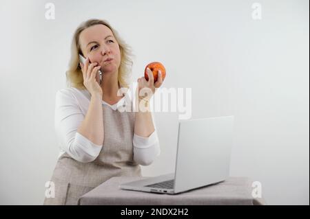 Femme au foyer dans un tablier beige à la maison fond blanc parler à des amis au téléphone manger de la pomme rouge et regarder des séries de films sur ordinateur portable insouciante vie calme à la maison confort de complaisance problème de potins Banque D'Images
